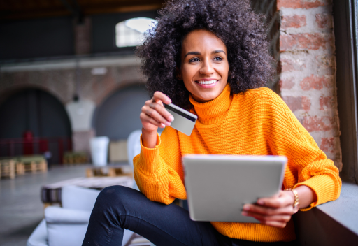 A smiling woman in an orange sweater holds a credit card in one hand and a table in the other, representing someone reading an email from a financial brand.